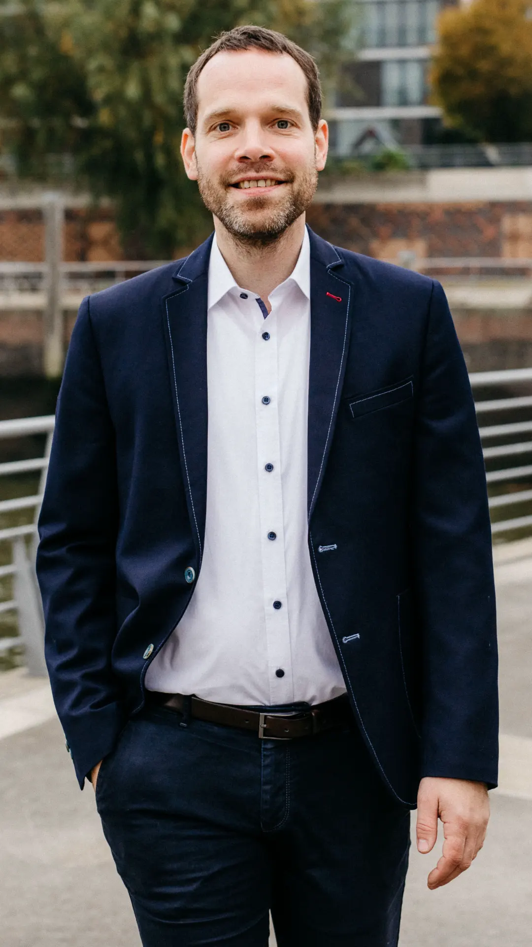 Das Bild zeigt Dennis Ahrens freundlich lächelnd und lässig mit seiner rechten Hand in der Hosentasche.